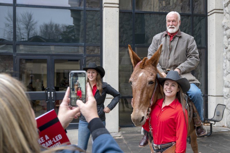 Missouri District 49 Representative Jim Schulte, New Bloomfield, climbs into the saddle atop UCM&rsquo;s live mule mascot, Tammy, also known as &ldquo;Mancow,&rdquo; during a visit to campus as part of the Legislative Freshman Tour. With him are student Mule Riders, Emma Lock, left, and Holly Hagood.