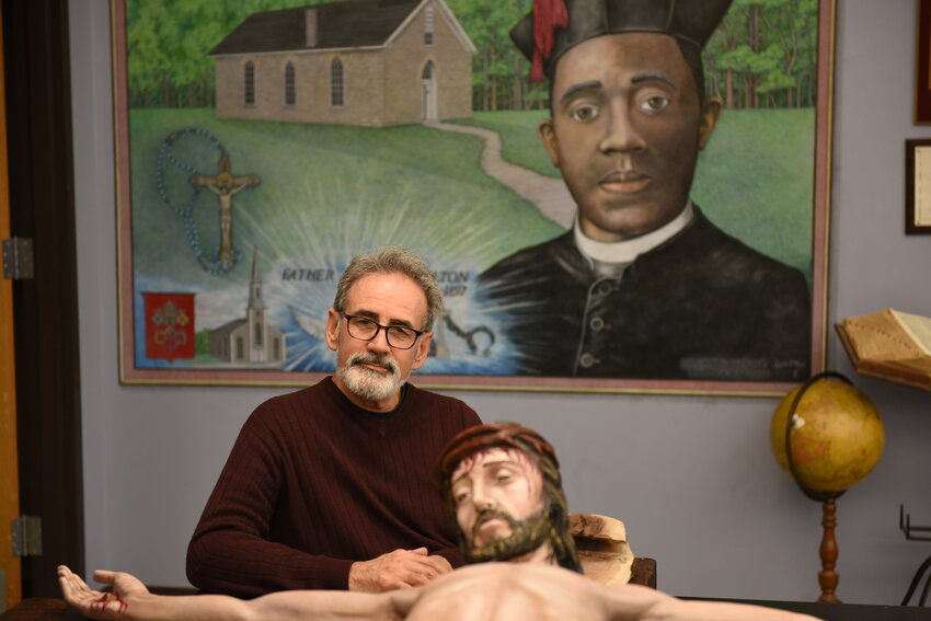 Art teacher Lonnie Tapia is seated next to the crucifix that now adorns the space above the sanctuary of the St. Thomas More Newman Center in Columbia. This was in march, while Mr. Tapia was restoring the crucifix in the Art Room of Fr. Tolton Regional Catholic High School in Columbia. The artist&rsquo;s &ldquo;Fr. Tolton Life Mural&rdquo; can be seen on the wall behind the crucifix.
