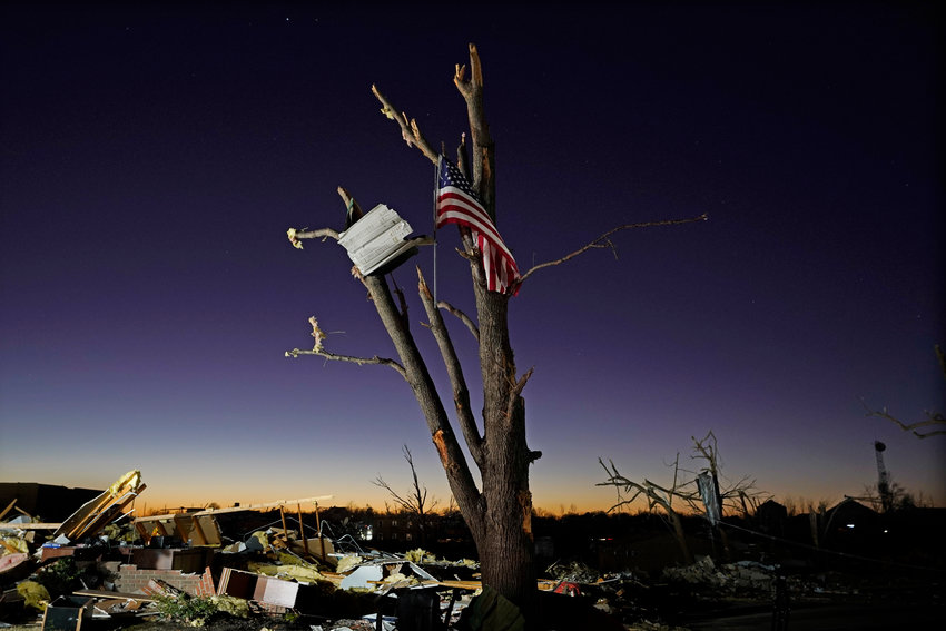 An American flag hangs from a damaged tree Sunday, Dec. 12, 2021, in Mayfield, Ky. President Joe Biden is headed to Kentucky to survey damage and offer federal support for the victims of the devastating tornadoes that killed dozens and left thousands more in the region without heat, water or electricity. More than 30 tornadoes tore through Kentucky and four other states over the weekend.  (AP Photo/Mark Humphrey)