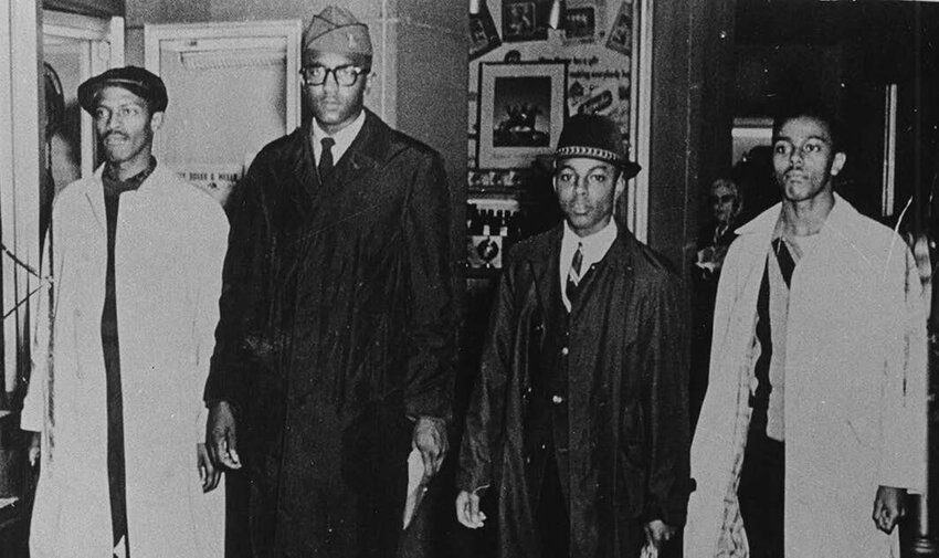 A stool on display at the NMAAHC. The picture in the background was taken on the second day of the Greensboro Lunch counter Sit-ins, Feb. 2, 1960. In the background photo above are A&T students Franklin McCain, Billy Smith and Clarence Henderson. In the original photo, Joseph McNeil is next to McCain on the far left.