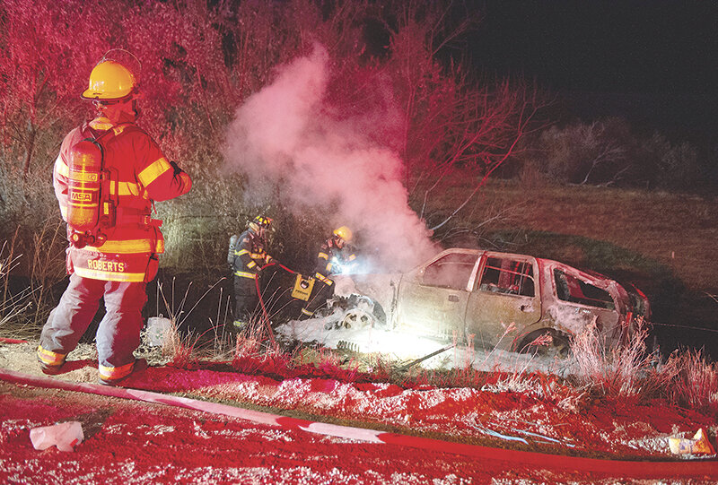 Powell firefighter Seth Roberts (at left) stands by the scene of a Sunday evening crash near Ralston, as two of his colleagues extinguish the vehicle fire. A woman and her two children were seriously injured in the crash.