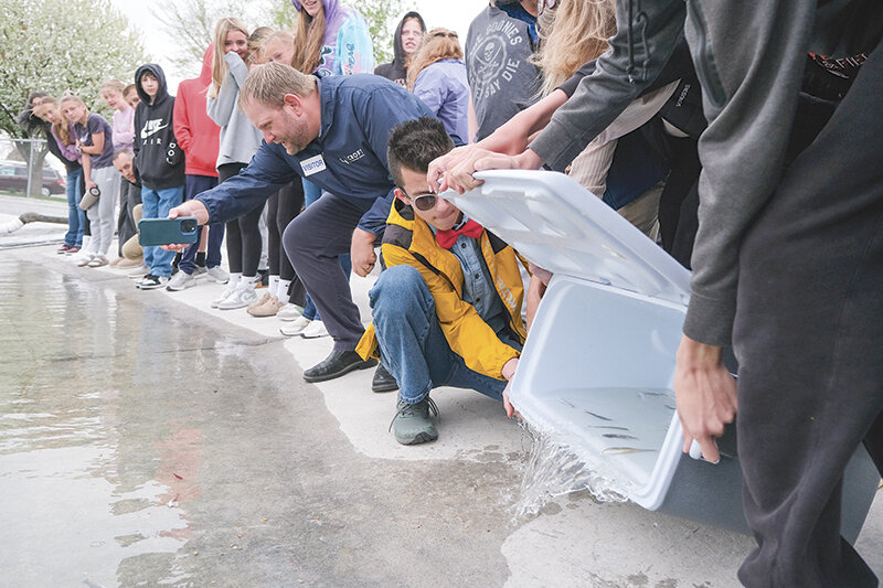 Powell Middle School student Noah MacDonald (in yellow) helps release the fish he and his class have cared for over the past five months into Homesteader Pond on May 5 while Patrick Croft (left) films the event and members of Powell Middle School&rsquo;s seventh and eighth grade classes watch carefully.