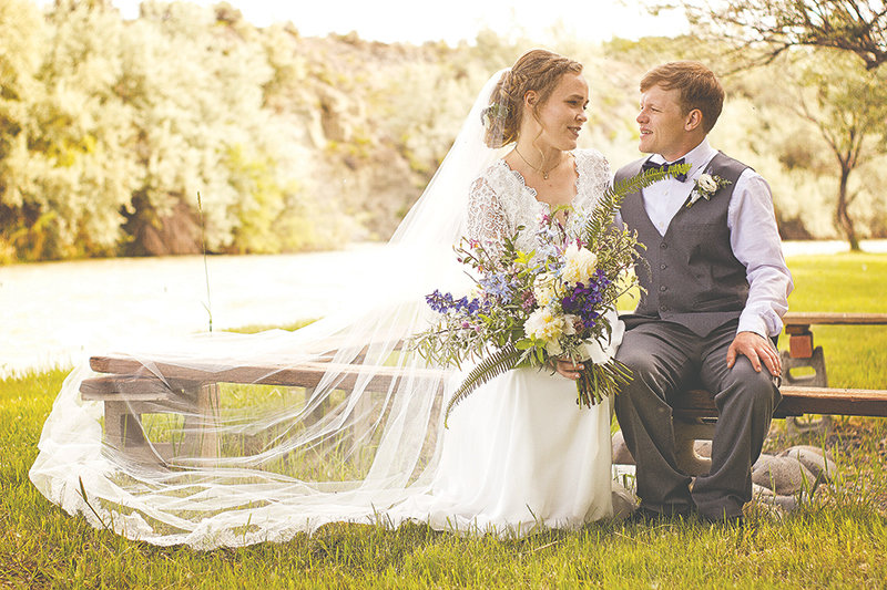 Bride and groom, Amanda and Colin Harnish, posed on their wedding day in June of 2020, seated on a bench in the riverside setting on the banks of the Shoshone River south of Powell.
