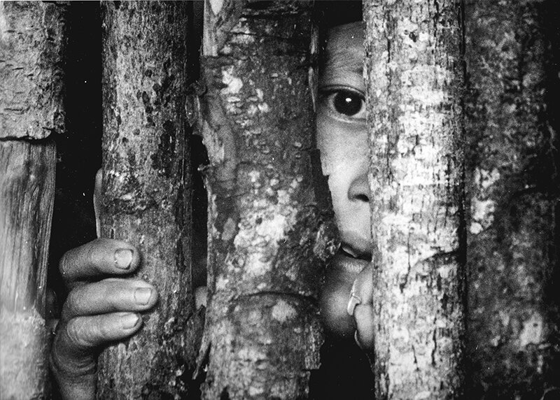 In 1987, a young member of an Ixil community in Guatemala’s Highland Mountains peers through the walls of his shelter made of sticks during the 30-year war in the Central American country.