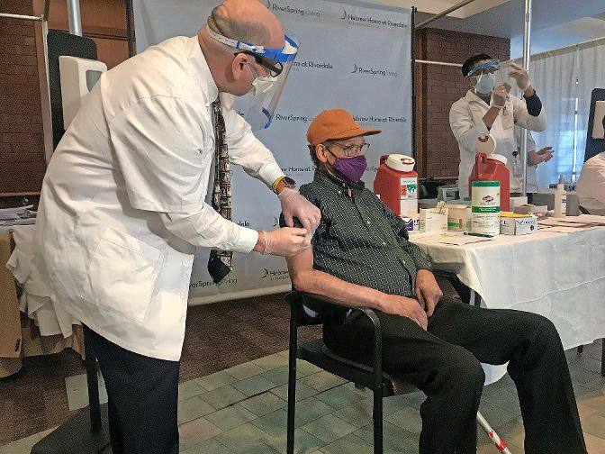 Kelley Dixon, 78, gets the first coronavirus vaccine among residents of the Hebrew Home at Riverdale on Monday morning. Walgreens pharmacists like Mitchel Zaretsky, left, and Ankur Amin administered inoculations to nearly 1,600 people at the Hebrew Home over the course of three days.