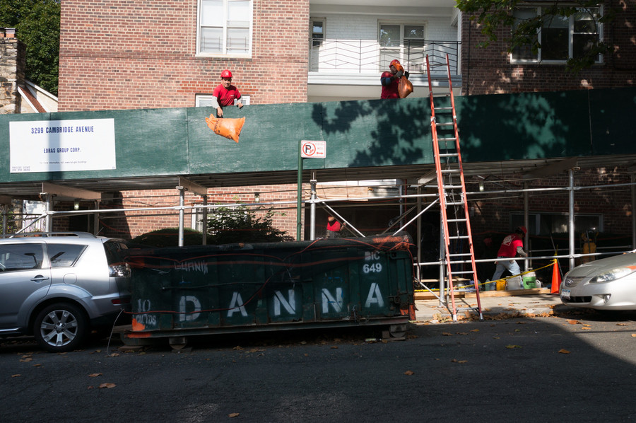 Construction workers atop scaffolding toss garbage into a dumpster.