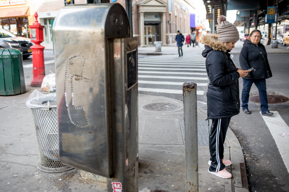 A pedestrian checks her phone while waiting at a crosswalk near a defunct pay phone at the corner of Broadway and West 231st Street. Some of the last few pay phones in Kingsbridge could be replaced by LinkNYC kiosks, which would bring free Wi-Fi, smartphone charging, a tablet to access city services, maps and directions to a handful of spots in the neighborhood — not just on Broadway, but also along Bailey Avenue.
