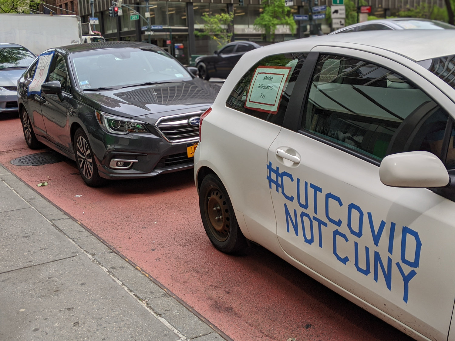 Blue tape pasted on the side of a car reads ‘cut COVID not CUNY’ alongside a sign reading ‘make billionaires pay’ during protests against layoffs at various schools within the CUNY system.
