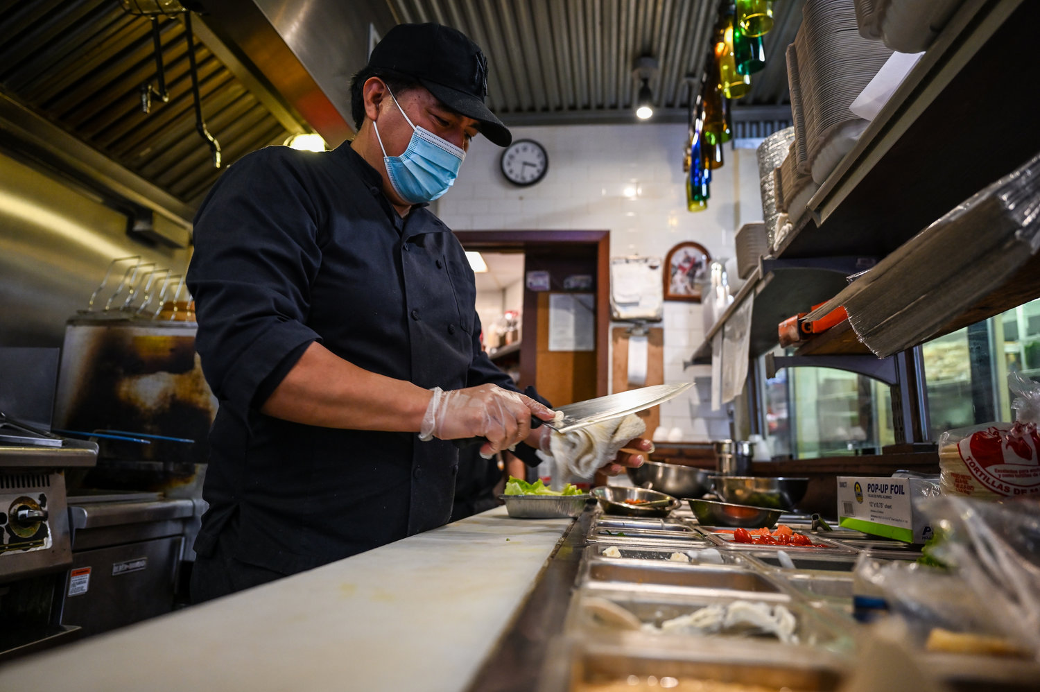 A cook at Taste & Sabor on West 231st Street and Riverdale Avenue cleans his knives during a lull in customers. While the eatery is known for its Latin and Greek fare, some neighbors in need have discovered that when Taste & Sabor receives more food than it needs, it’s willing to share.