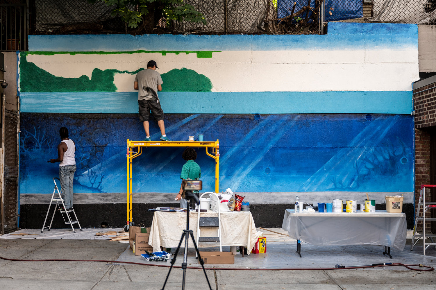 Artist Nicky Enright joins a volunteer painting a mural on the back retaining wall of 3636 Greystone Ave., where he lives. Enright was commissioned to paint this mural, ‘Aquarium,’ by his building’s co-op board because he’s no stranger to mural painting.