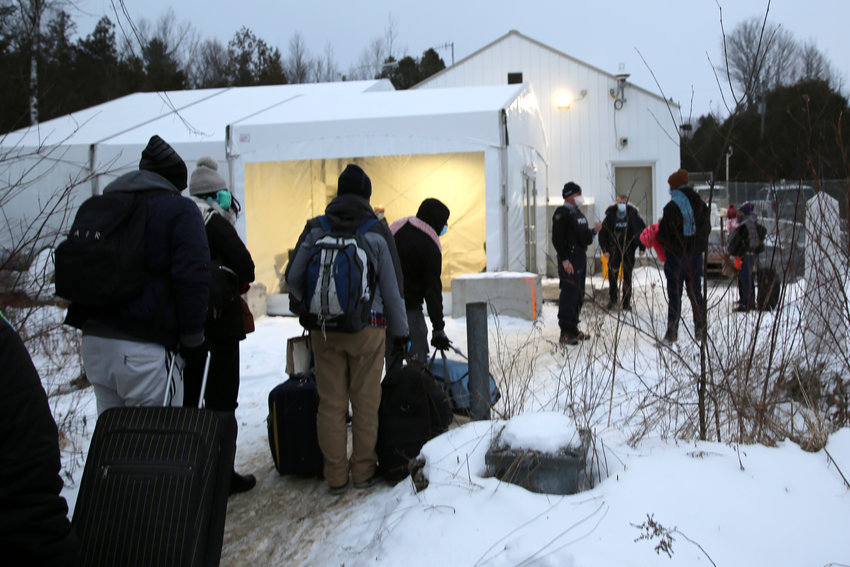 HOLD FOR STORY BY WILSON RING &mdash; Migrants line up on the border of United States, foreground, and Canada, background, at a reception center for irregular borders crossers, in Saint-Bernard-de-Lacolle, Quebec, Canada, Wednesday Jan. 12, 2022, in a photo taken from Champlain, N.Y. They are crossing the U.S.-Canadian border into Saint-Bernard-de-Lacolle, Quebec, where they are arrested by the Royal Canadian Mounted Police and then allowed to make asylum claims. The process was halted for most cases after the 2020 outbreak of COVID-19, but the Canadian government changed its policy in November, allowing the process to continue.