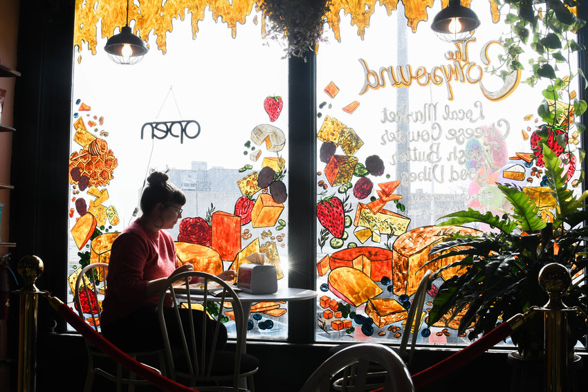 WINDOW SEAT &mdash; A woman eats in front of a painted window featuring different types of cheese at The Compound in Utica.