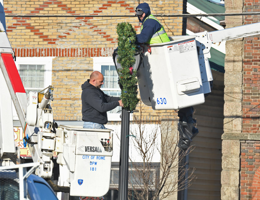 GETTING A JUMP ON THE HOLIDAYS &mdash;&nbsp;City of Rome workers Larry Fisher, left, and Lenny Costello each get a lift in a bucket truck to the top of one of the city&rsquo;s decorative light poles as they install colorful holiday wreaths along East Dominick Street on Wednesday. Over the course of the next few weeks, city workers will install dozens of holiday pole decorations, trim the city&rsquo;s holiday tree and install the whimsical Trinkaus holiday village and iconic Merry Christmas sign on North James Street. It will be the first holiday season that the popular Trinkaus sign and display will be lit since the death of its creator, Andy Trinkaus, who passed away at age 99 on July 14.
