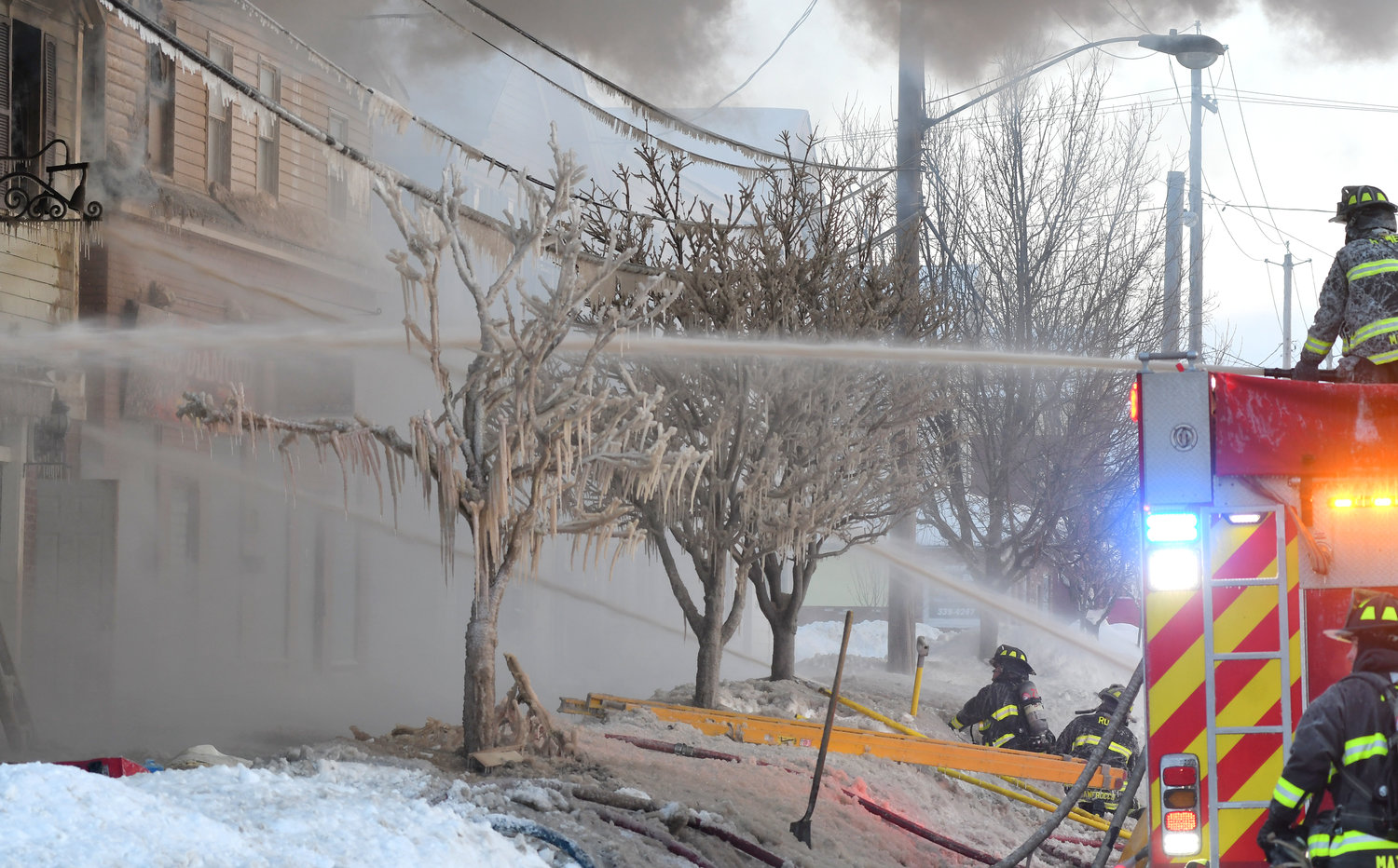 STILL WORKING — Firefighters battle the blaze at 305 N. James St. this morning, dousing flames as icycles hang from trees in front of the building and smoke billows into the morning sky.