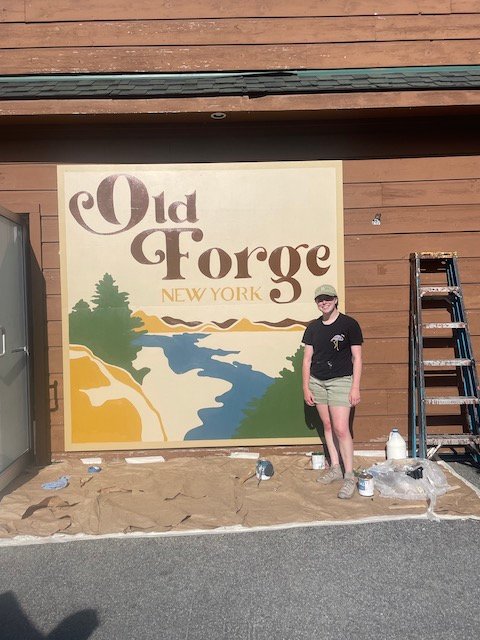 Char Miller, a summer intern at  View, in front of the mural she created at Souvenir Village by the five corners/Point Park in Old Forge.