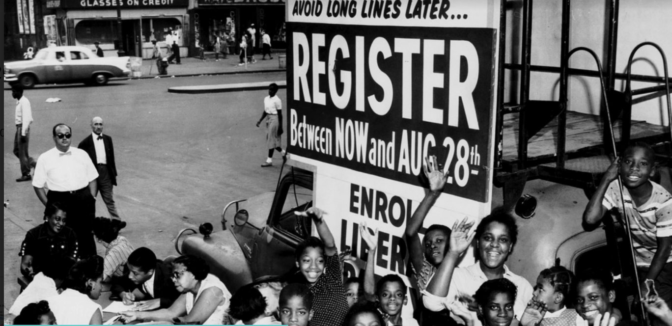A group of African-American children gather around a sign and booth to register voters during the 1960s.