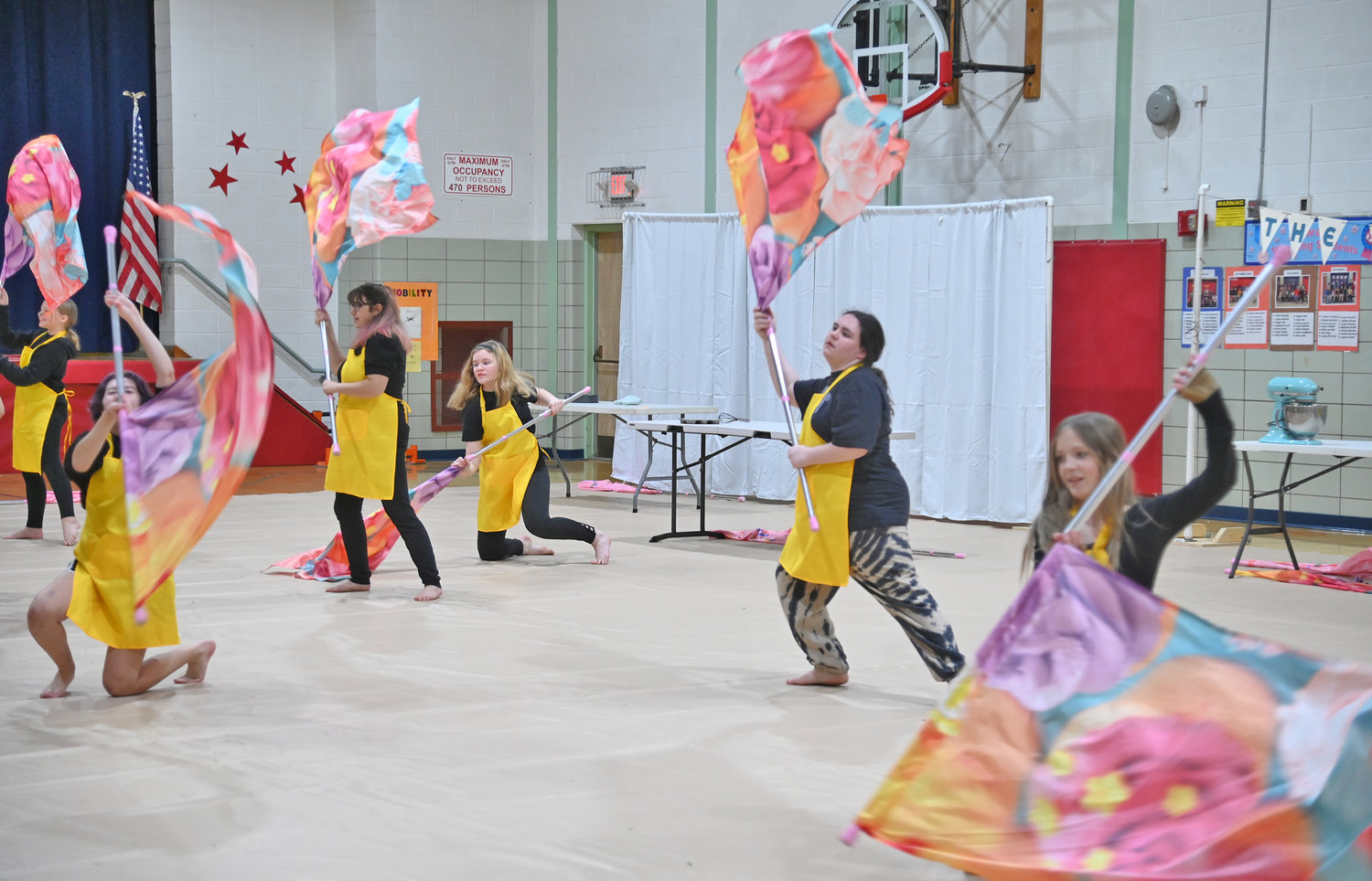 Members of the New Hartford JV Winter Guard go through the paces of their show called “Junior Bake-Off” about a baking competition. The guard members wear bright yellow aprons and spin brightly-colored flags, with cupcakes and rainbow sprinkles on them.