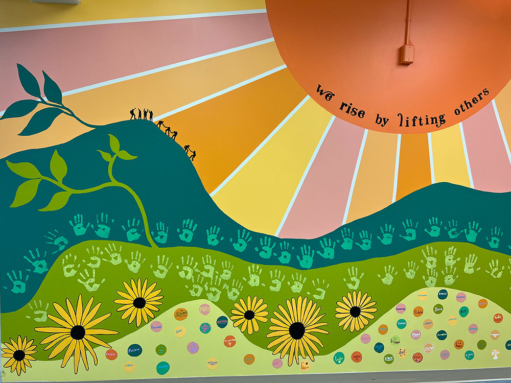 On the far left of the mural, students can be seen giving one another a hand to help everyone rise up a hill. A radiant sun was added to the center, and on the right is an oak tree. Across the rolling hills are the handprints of each of the 120 rising middle school students, and oversized black-eyed Susans.